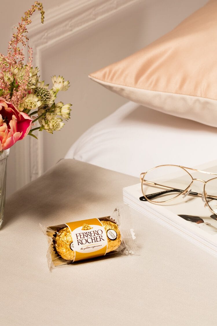 Fotografía profesional de dos Ferrero Rocher sobre una mesita blanca junto a un jarrón con flores y unas gafas en entorno elegante