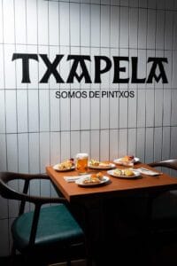 Mesa con cinco mejores tapas españolas en restaurante Txapela Madrid, muro con nombre y slogan “Somos de pintxos” al fondo, fotografía gastronómica profesional