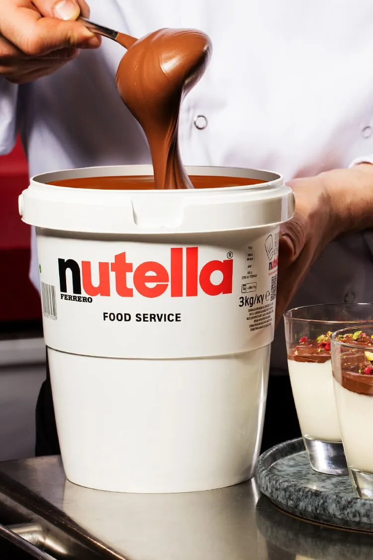Chef dejando caer Nutella con una cuchara sobre el bote original, junto a copas de postre, fotografía profesional de producto en España