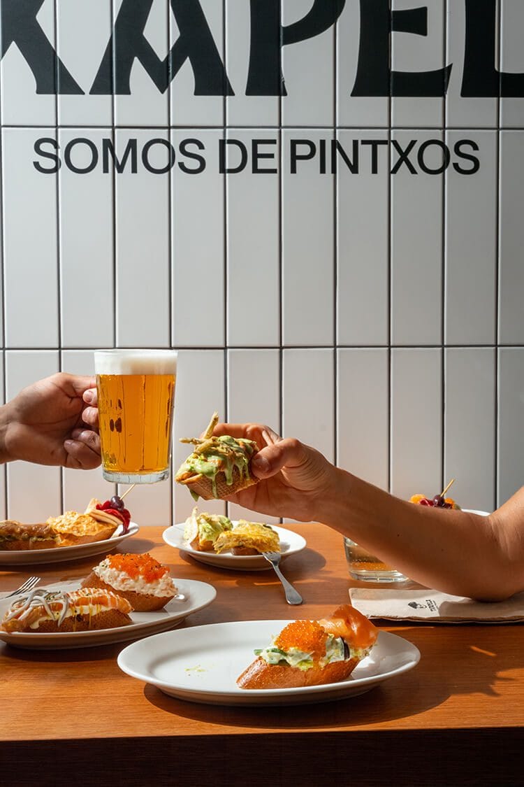 Clientes disfrutando tapas y cerveza con el mural de Txapela y el lema “Somos de Pintxos” al fondo en Madrid.