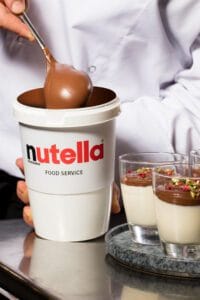 Bote de Nutella en primer plano con cucharón cayendo y copas de postre gourmet al fondo, fotógrafo profesional de publicidad y productos en España.