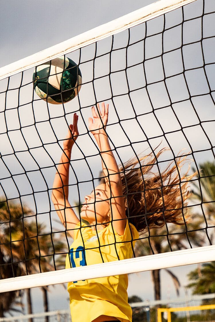 Fotografía profesional de retrato deportivo en Barcelona de una chica jugando al volley playa, saltando hacia la red con el cielo y el mar de fondo.