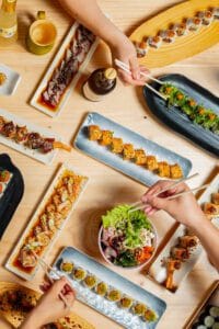 Mesa decorada con variedad de sushi y manos usando palillos para coger comida en el restaurante Bodegón Sushi Kurama, fotógrafo profesional en Barcelona, España.