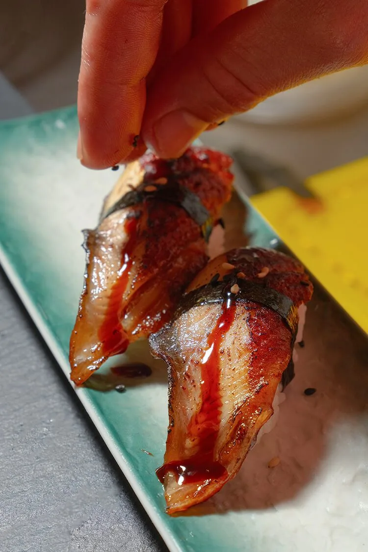 Nigiris de anguila flambeada decorados con salsa y semillas de sésamo, con mano espolvoreando el sésamo en Bodegón Sushi Kurama, fotógrafo profesional en Barcelona.