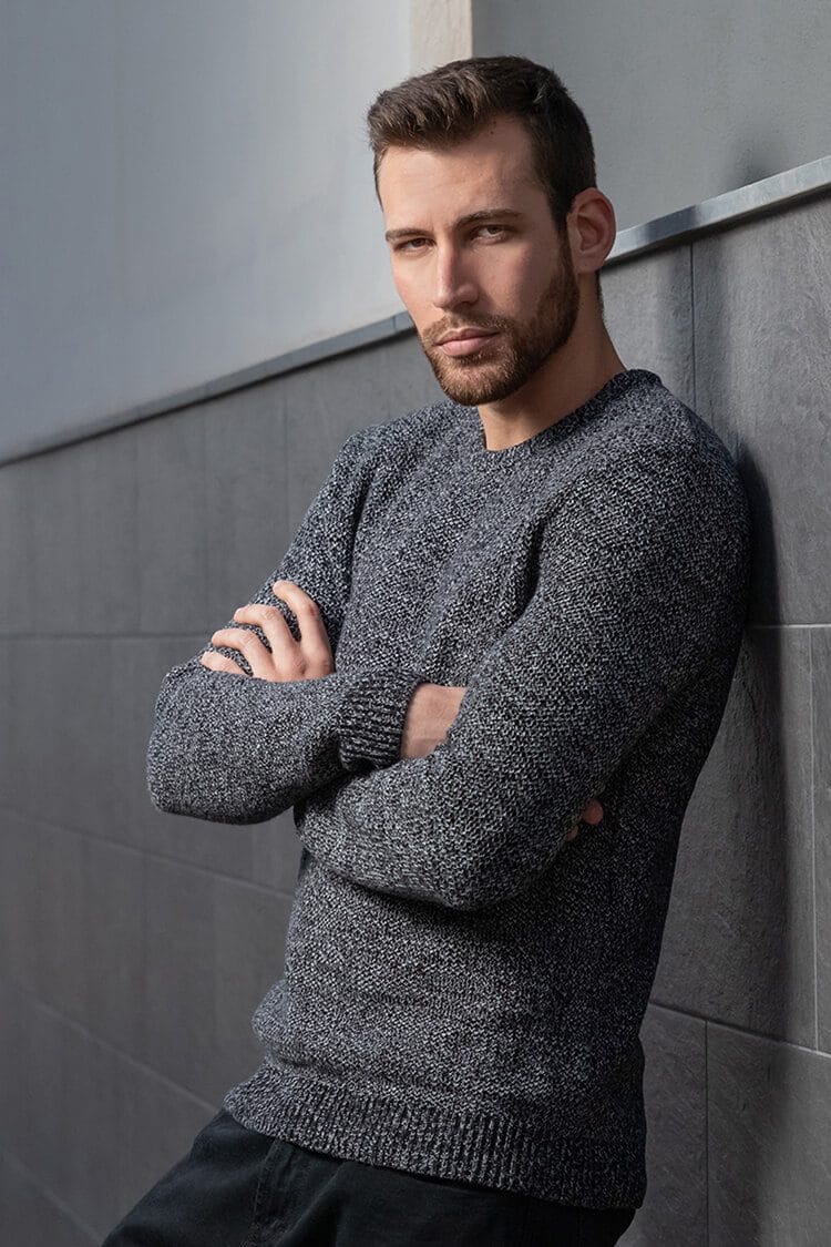 Retrato de Jadier con sonrisa sensual, brazos cruzados y apoyado en la pared en una pose informal y moderna.