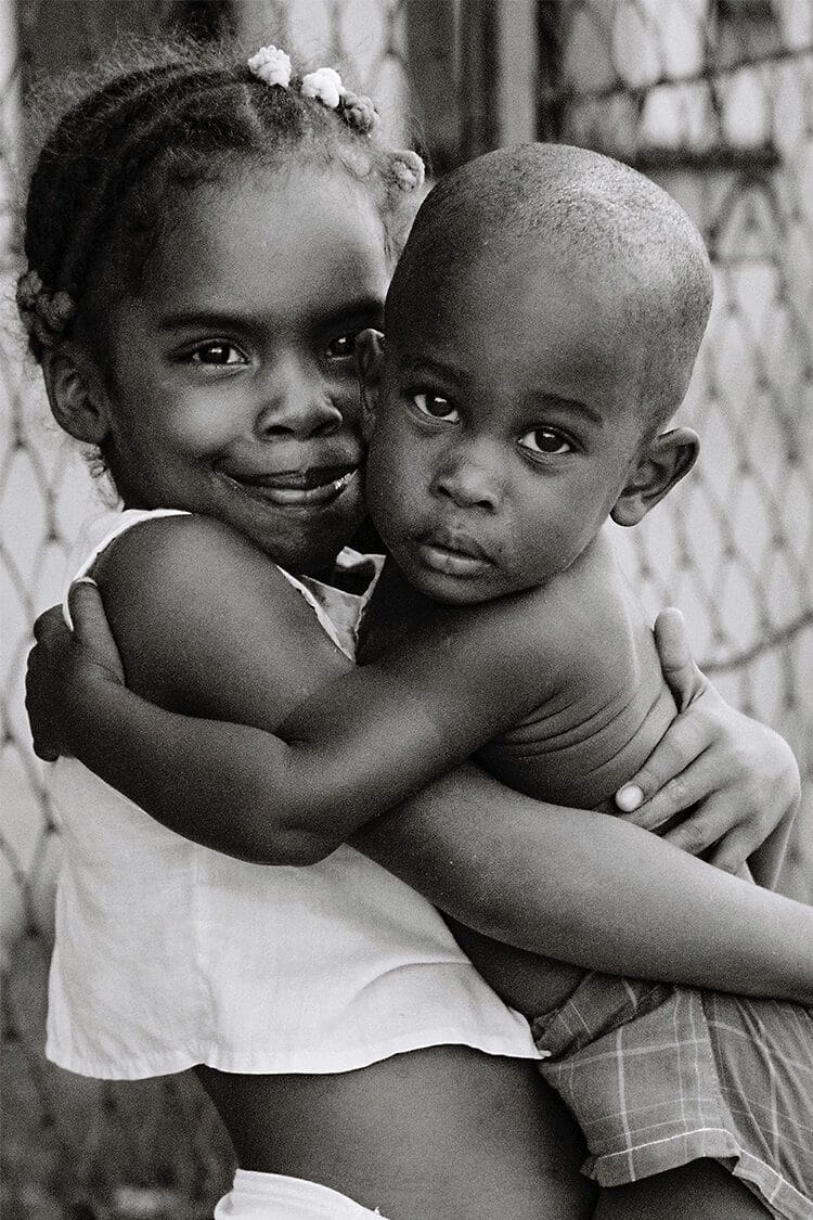 Retrato emocional en blanco y negro de una niña cubana abrazando con ternura a su hermano bebé, sonriendo con alegría.