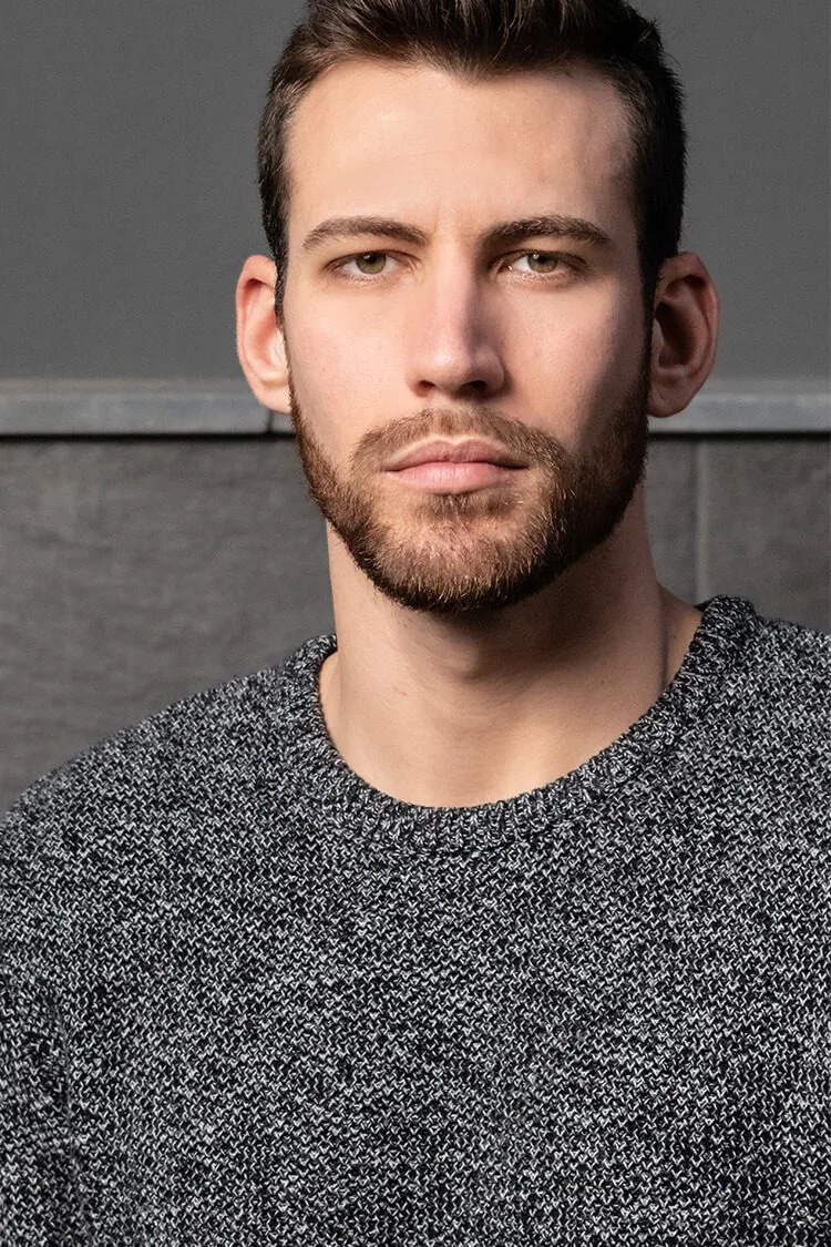 Retrato masculino profesional de Jadier serio, con jersey gris, barba completa y mirada intensa en estudio.