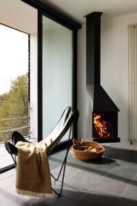 Habitación con chimenea vertical en la pared, una silla en salón y terraza con cristalera corredera abierta