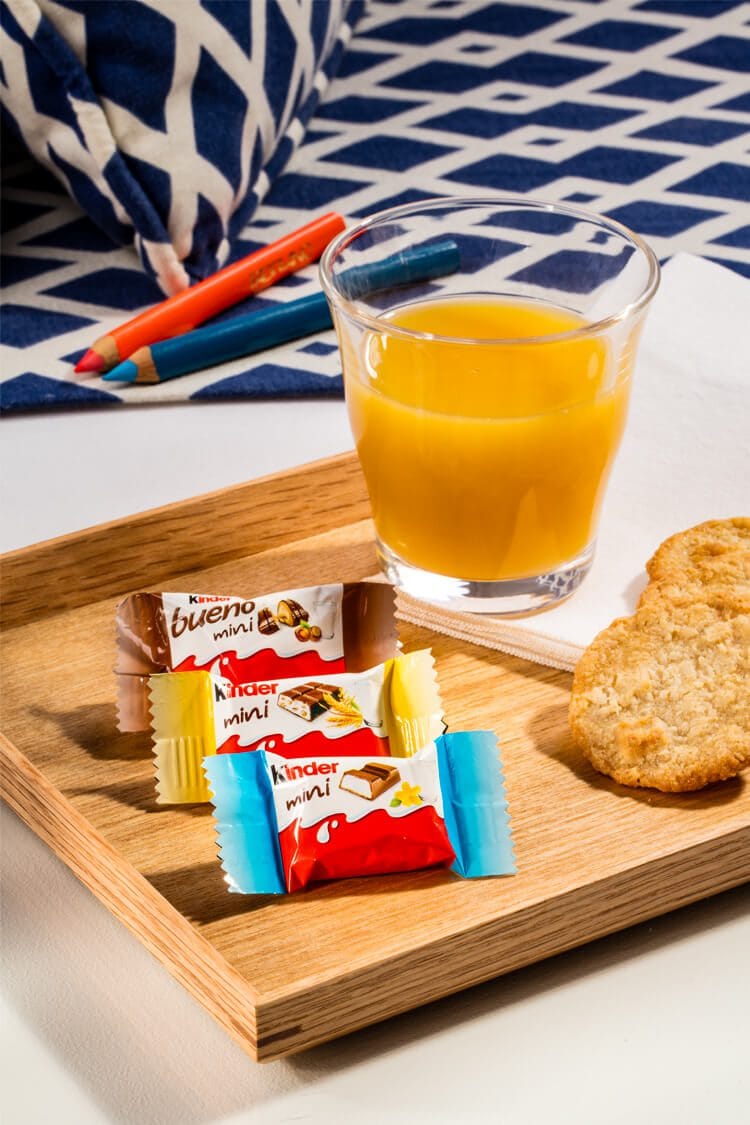 Fotografía profesional de tres paquetes de mini chocolates Kinder junto a una tostada y un zumo de naranja en un desayuno continental