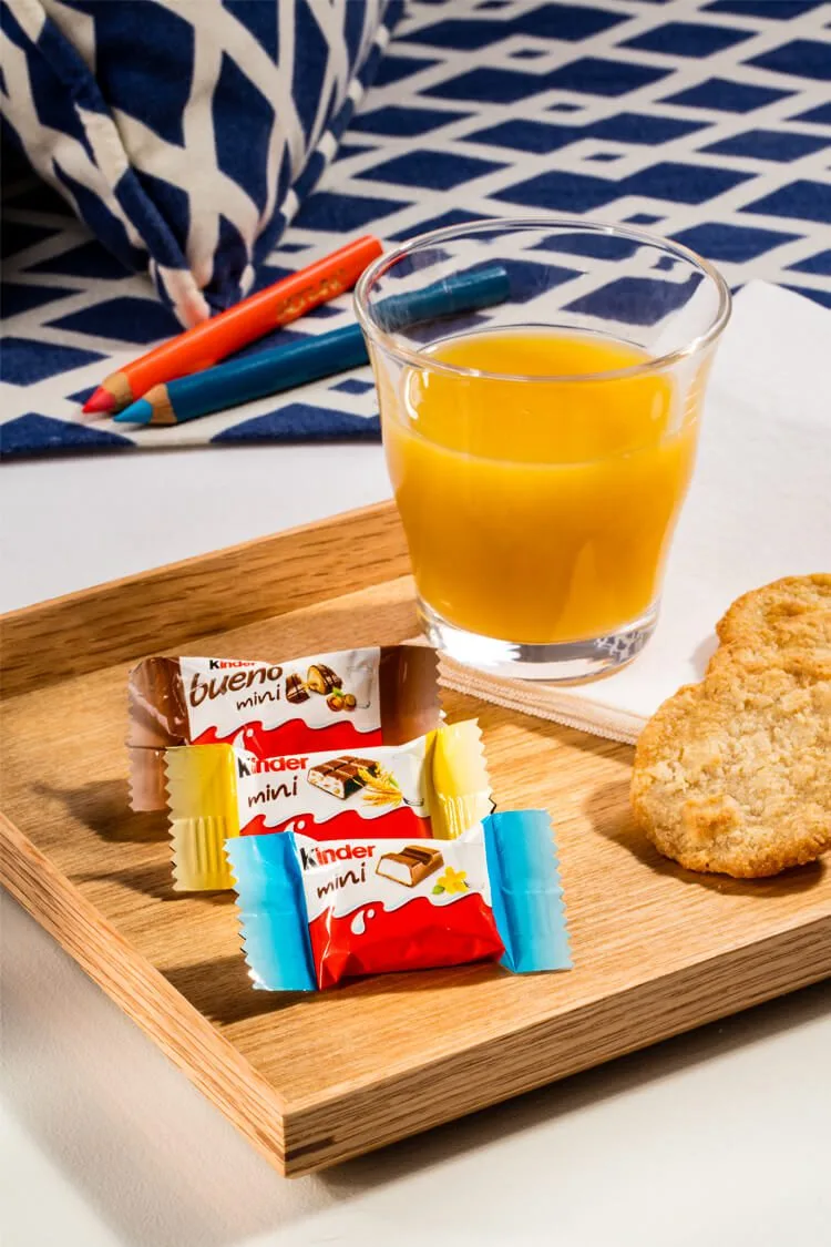 Fotografía profesional de tres paquetes de mini chocolates Kinder junto a una tostada y un zumo de naranja en un desayuno continental