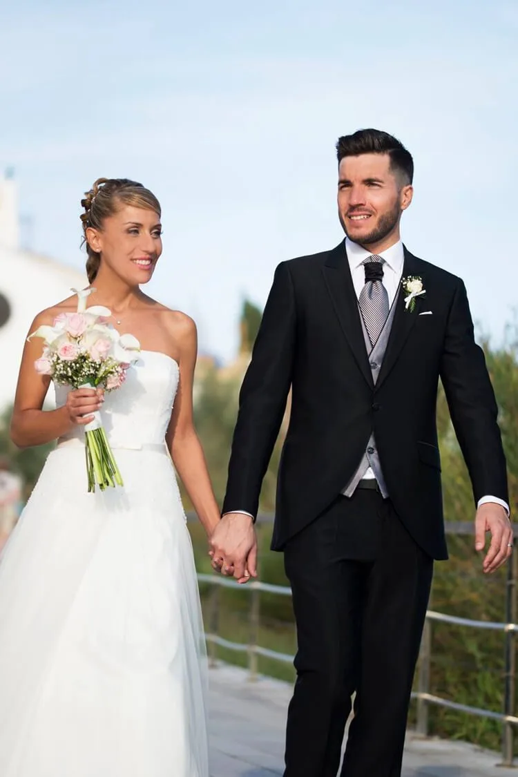 Pareja caminando cogidos de la mano por la costa de Barcelona, novia con ramo de flores