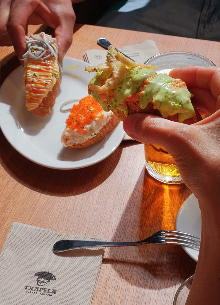 Plato de pinchos fusión en el centro de la mesa con manos de clientes cogiéndolos en el restaurante Txapela de Madrid.