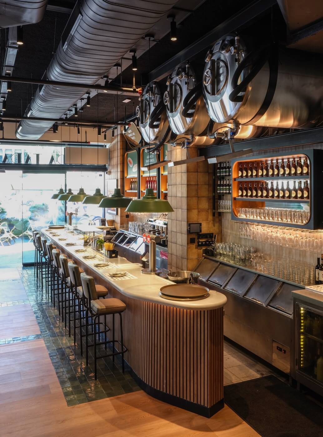 Imagen vertical de la barra del restaurante Tapa Tapa desde el lado derecho, con lámparas verdes, barriles y estanterías de cervezas artesanas.