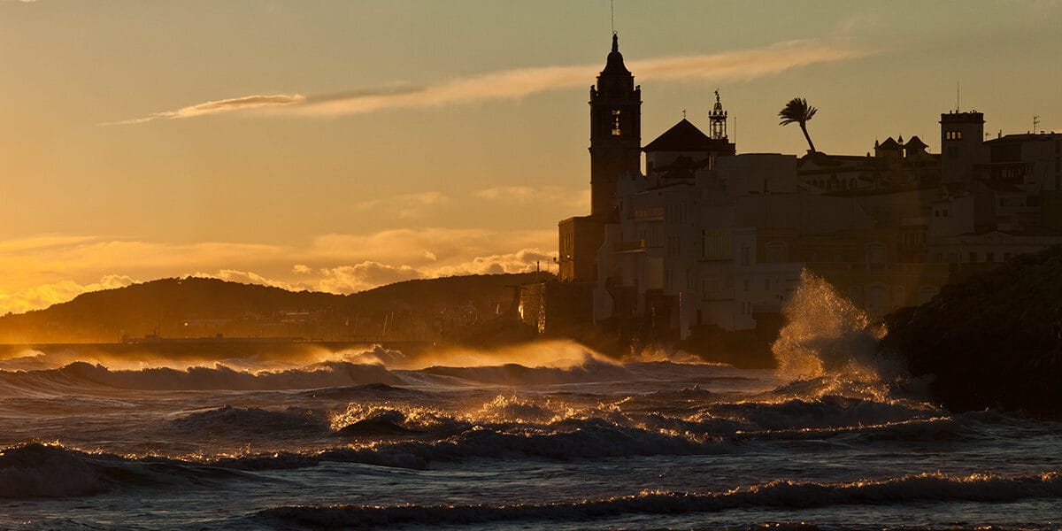 Olas rompiendo junto a la playa de Balmins en Sitges al atardecer, con la silueta de la iglesia de Sant Bartomeu al fondo, fotografía de paisaje en Barcelona