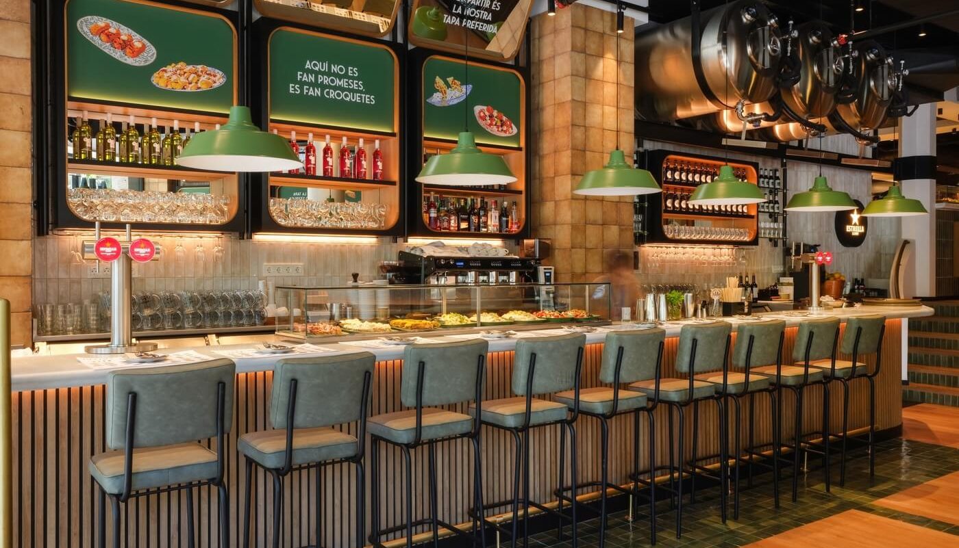 Fotografía profesional de la barra principal del restaurante Tapa Tapa en Barcelona con iluminación cálida, taburetes alineados y surtidor de cerveza Estrella Damm.