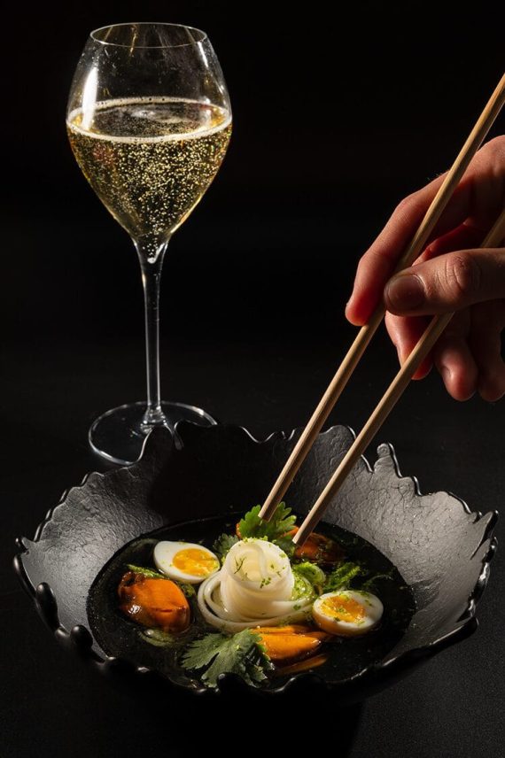 Plato gourmet de ramen con huevo, zanahoria y copa de vino blanco en el restaurante Sensacions de Barcelona, fotografía gastronómica profesional