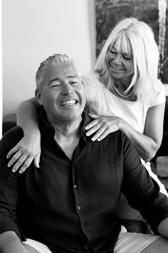 Mujer abrazando por los hombros a su pareja sentada, ambos riendo, retrato familiar en blanco y negro